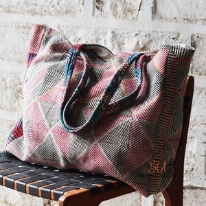 May include: A large tote bag featuring a quilted texture with a geometric pattern in shades of red, white, and black. The bag has two matching fabric handles and displays the text BUI130 in the top right corner and BU in the bottom right.