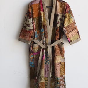 May include: A knee-length robe featuring a patchwork design of various fabric squares in shades of brown, tan, pink, and blue. The garment includes a solid tan trim along the front opening, three-quarter length sleeves, and a matching tie belt.