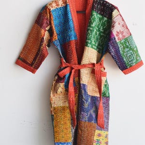 May include: A patchwork robe featuring a variety of colorful fabric squares in shades of blue, green, yellow, and brown. The garment has three-quarter length sleeves, a solid orange trim along the edges, and a matching fabric tie belt at the waist.