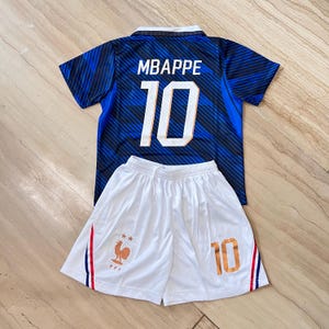 Può includere: Una maglia da calcio a motivi blu e neri con il nome MBAPPE e il numero 10 stampati in bianco sul retro, abbinata a pantaloncini da calcio bianchi con un emblema di gallo dorato, due stelle e il numero 10 in oro sulla gamba anteriore.