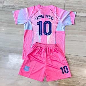 May include: A pink and light blue sports jersey and shorts set. The jersey features the name LAMINE YAMAL and the number 10 in dark blue on the back. The matching pink shorts display the number 10 and a small blue and red team crest emblem.