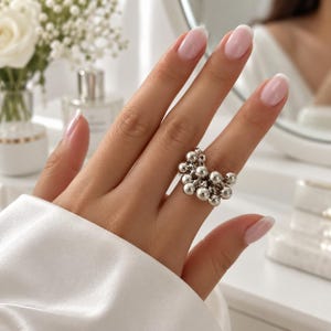 Puede incluir: Un anillo de tono plateado que presenta un grupo de múltiples cuentas esféricas pequeñas y lisas, dispuestas en una formación densa y redondeada. El anillo se lleva en un dedo, mostrando la superficie metálica y reflectante de las esferas.