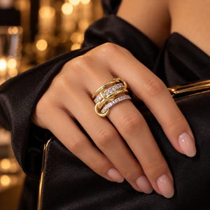 May include: A multi-layered ring featuring interlocking gold-toned bands, with two of the bands encrusted with small, clear, sparkling stones. The design creates a wide, textured appearance across the finger, combining smooth polished metal with rows of brilliant, light-reflecting crystals in a cohesive, layered arrangement.