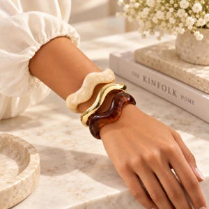 Puede incluir: Un conjunto de tres brazaletes ondulados que se llevan en la muñeca. El conjunto incluye un brazalete de color crema con efecto mármol, un brazalete de metal dorado pulido y un brazalete de color marrón oscuro con patrón de carey.