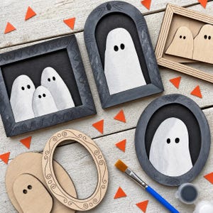 Puede incluir: Una colección de decoraciones de pared de madera con temática de fantasmas, con varios marcos en acabados negro y madera natural. Los fantasmas son blancos con ojos negros. Los marcos son rectangulares, arqueados u ovalados, con un pincel y pintura.