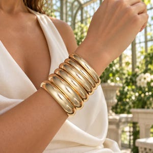 Może przedstawiać: Stos sześciu polerowanych, złotych metalowych bransoletek typu bangle noszonych na nadgarstku. Każda bransoletka ma gładką, zaokrągloną i odblaskową powierzchnię o ciągłym, okrągłym kształcie. Bransoletki są jednolite pod względem rozmiaru i koloru.