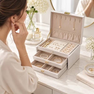 Puede incluir: Un joyero blanco de varios niveles con tapa abatible, interior forrado en terciopelo con ganchos para collares, una bandeja para pendientes y dos cajones extraíbles para organizar anillos, pulseras y relojes con herrajes dorados.