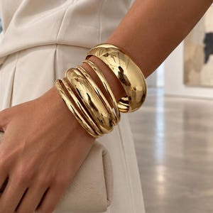 May include: A set of five polished gold-tone metal bangles of varying thicknesses worn together. The bracelets feature a smooth, reflective surface with a rounded profile, creating a stacked appearance on the wrist. The metal has a bright yellow gold finish.