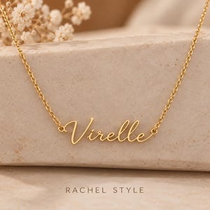 Puede incluir: Un collar de tono dorado con una cadena fina y un colgante central de placa con el texto Virelle en letra cursiva. Debajo del colgante, el texto RACHEL STYLE está impreso en una fuente sans-serif sobre la superficie de apoyo.
