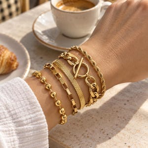 Puede incluir: Un conjunto de cinco pulseras doradas usadas juntas. La colección incluye una cadena de cuentas, una cadena de eslabones rectangulares, una cadena de espiga plana, una cadena de eslabones gruesos con cierre de barra y una cadena de cuerda trenzada.