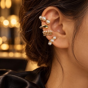 Puede incluir: Un conjunto de cuatro pendientes tipo ear cuff dorados colocados en el cartílago y el lóbulo. El superior tiene dos perlas blancas, el segundo es una banda dorada lisa, el tercero tiene una fila de perlas pequeñas y el inferior muestra tres perlas blancas.