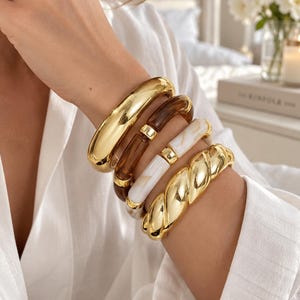 Puede incluir: Un conjunto de cuatro pulseras que incluye un brazalete dorado liso, uno de resina marrón oscuro, uno de resina blanca veteada y un brazalete de metal dorado trenzado. Las pulseras se llevan juntas en la muñeca, mostrando texturas y acabados.