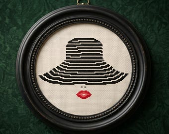 May include: A cross-stitch embroidery piece framed in a round black frame. The design features a wide-brimmed hat rendered in horizontal black and white stripes, positioned above a pair of bright red lips on a cream-colored fabric background.