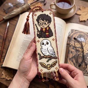 May include: A rectangular cross-stitch bookmark on cream fabric featuring a cartoon boy with glasses and a wand, a white owl on a branch, and a golden winged ball. The bookmark has a braided tassel at the top and beaded fringe at the bottom.