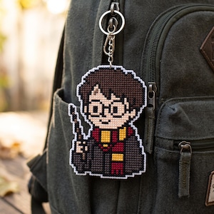 May include: A pixel art style keychain featuring a character with dark brown hair, round glasses, a black robe, and a red and yellow striped scarf, holding a wand. The figure is crafted with a grid-like texture and attached to a silver metal ring.