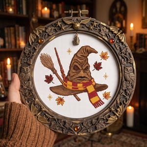 May include: A circular embroidery hoop frame with an ornate bronze-colored border featuring owls, leaves, and wings. Inside, a cross-stitch design depicts a brown sorting hat, a broomstick, a wand, and autumn leaves on white fabric.