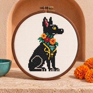 Puede incluir: Un bastidor de bordado circular de madera que contiene un patrón de punto de cruz de un perro negro sentado de perfil. El perro está adornado con un collar floral colorido con hilos naranjas, rojos y verdes, e incluye detalles amarillos.