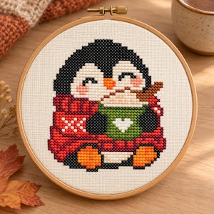 May include: A circular wooden embroidery hoop containing a cross-stitch design of a penguin with black, white, and orange features. The penguin wears a red patterned scarf and holds a green mug with a white heart, filled with a brown beverage.
