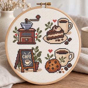 May include: A circular wooden embroidery hoop containing a cross-stitch pattern on white fabric. The design features four coffee-themed motifs: a brown coffee grinder, a slice of chocolate cake with a mug, a cafe sign, and a cookie with a mug.