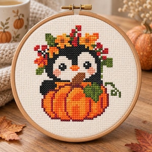 May include: A circular wooden embroidery hoop containing a cross-stitch pattern of a black and white penguin wearing a crown of orange flowers and red berries, sitting behind a large orange pumpkin with a green leaf and vine on a white fabric.