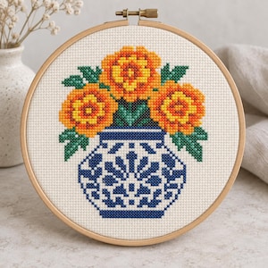 May include: A circular wooden embroidery hoop containing a cross-stitch pattern of three orange and yellow flowers in a blue and white patterned vase. The design is stitched onto white Aida fabric, featuring green leaves and a detailed geometric base.