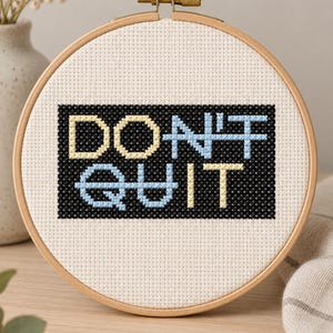 May include: A circular wooden embroidery hoop containing white Aida fabric with a cross-stitch design. The design features a black rectangular background with the text DON'T QUIT stitched in alternating light blue and pale yellow thread colours.