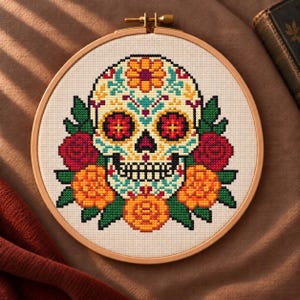 Può includere: Un telaio da ricamo circolare in legno contenente un motivo a punto croce di un teschio decorato. Il teschio è ornato da motivi floreali colorati nelle tonalità dell'arancione, rosso, giallo e turchese, circondato da foglie verdi su tessuto bianco.