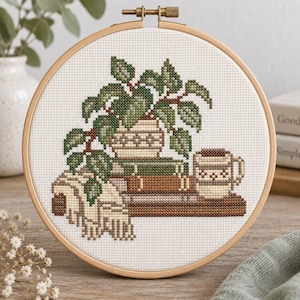 May include: A circular wooden embroidery hoop containing a cross-stitch pattern on white fabric. The design features a potted green plant, a stack of three books in shades of green and brown, a beige mug, and a draped fabric with fringe detail.