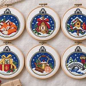 May include: Six circular wooden embroidery hoops containing cross-stitch designs of Christmas ornaments. The patterns feature a sleeping fox, a lantern with a red bow, a birdhouse with a cardinal, wrapped gifts, a teapot with cinnamon, and a stone bridge.
