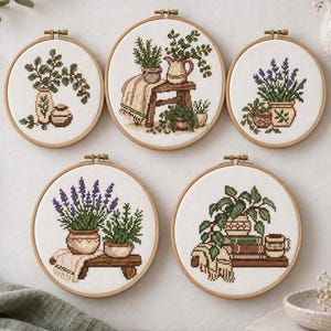 Könnte beinhalten: Fünf runde Stickrahmen aus Holz mit Kreuzstichmotiven von Topfpflanzen, Kräutern und Blumen. Die Muster zeigen botanische Arrangements, Keramikvasen, Holzbänke und gefaltete Textilien in Grün-, Lila- und Brauntönen auf weißem Stoff.