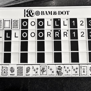 May include: A white rectangular Mahjong score card with the text BAM & DOT at the top. The board features a grid with black and white tiles, including letters, numbers, and symbols. A row of tiles with floral, dragon, and directional designs sits below.