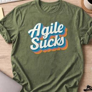 Puede incluir: Una camiseta de manga corta de color verde oliva con el texto "Agile SUCKS" impreso en una fuente blanca y llamativa de estilo retro. Las letras están superpuestas con varias sombras paralelas de colores en tonos azul, morado, rosa y naranja.