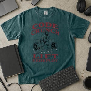 May include: A dark teal short-sleeve t-shirt featuring a graphic of a person lifting a barbell. The text reads CODE CRUNCH LIFT REDEPLOY in red lettering. The design includes illustrations of weight plates and gym equipment.