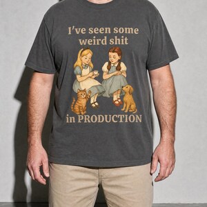Op de afbeelding: Een antracietgrijs T-shirt met korte mouwen en een afbeelding van twee meisjes, een kat en een hond. De tekst luidt I've seen some weird shit in PRODUCTION. De illustratie toont twee meisjes in jurken, waarvan er één een theekopje vasthoudt, met een kat en een hond.