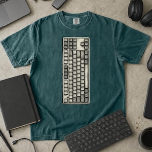 May include: A dark teal short-sleeve t-shirt featuring a central graphic of a vintage, weathered computer keyboard with off-white keys and a worn texture. The shirt has a crew neckline and a relaxed fit, showcasing a detailed print of keys.