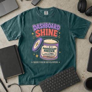 Może przedstawiać: Ciemnozielony t-shirt z krótkim rękawem z grafiką fioletowego pojemnika z napisem DASHBOARD SHINE. Tekst głosi DASHBOARD SHINE, makes questionable metrics look executive-ready oraz MADE FOR BI DEVELOPERS.