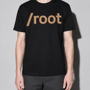 Puede incluir: Una camiseta negra de manga corta con el texto /root impreso en un estilo de letras de bloque tridimensionales de color beige y marrón en el pecho. La prenda tiene un cuello redondo clásico y un dobladillo recto, ofreciendo una silueta simple.