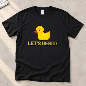 May include: A black short-sleeve t-shirt featuring a pixel art graphic of a yellow rubber duck with an orange beak. Below the duck, the text LET'S DEBUG is printed in a yellow pixelated font. The shirt has a crew neckline and a standard fit.