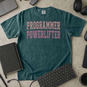 May include: A dark teal short-sleeve t-shirt with a crew neckline. The centre of the shirt features the text "PROGRAMMER POWERLIFTER" printed in a light pink, distressed, block-style font. The fabric has a washed, vintage appearance.