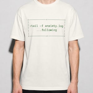 Puede incluir: Una camiseta de manga corta color blanco roto con un gráfico rectangular que contiene el texto en verde oscuro >tail -f anxiety.log y ...following. El diseño incluye finas líneas negras que forman un borde alrededor del texto.
