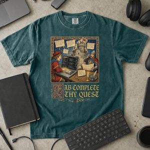 Può includere: Una t-shirt a maniche corte color verde acqua scuro con un'illustrazione medievale che ritrae una persona che usa un portatile in un castello. Il testo "TAB COMPLETE THY QUEST" è stampato sotto in un carattere decorativo color oro.