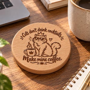 May include: A round light-coloured wooden coaster featuring a laser-engraved illustration of a fluffy cat wearing sunglasses and a crown, next to a steaming mug. The text reads: Cats don't drink cocktails, Make mine coffee, with small crown accents.