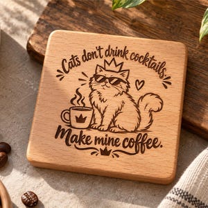 Op de afbeelding: Een vierkante houten onderzetter met een lasergegraveerde illustratie van een kat met een zonnebril en een kroon, naast een dampende koffiemok. De tekst luidt: "Cats don't drink cocktails, make mine coffee." Het ontwerp bevat kleine hartjes en kroontjes.
