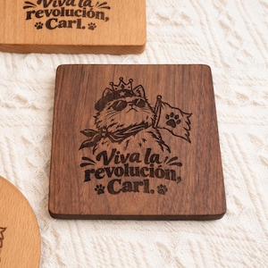 May include: A square dark wood coaster featuring a laser-engraved illustration of a cat wearing a beret, sunglasses, and a crown, holding a flag with a paw print. Below the cat, the text reads: Viva la revolución, Carl. with paw prints.
