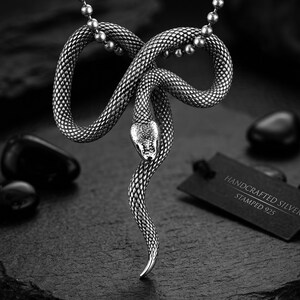 Puede incluir: Un colgante de serpiente en tono plateado suspendido de una cadena de bolas. La serpiente presenta texturas de escamas detalladas y una cabeza lisa. Una pequeña etiqueta rectangular negra muestra el texto HANDCRAFTED SILVER STAMPED 925.