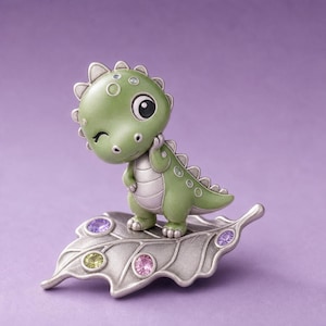 Puede incluir: Una pequeña figura de dinosaurio verde con expresión de guiño, de pie sobre una base de hoja de metal plateado. La hoja está decorada con tres piedras preciosas redondas y facetadas en colores púrpura, rosa y verde claro incrustadas.