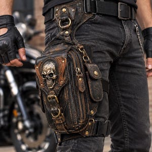 Può includere: Una borsa da gamba in pelle marrone e nera dall'aspetto vissuto, caratterizzata da un emblema metallico a forma di teschio sulla patta frontale, scomparti multipli con cerniera, finiture in ottone e cinghie in nylon regolabili per la vita e la coscia.