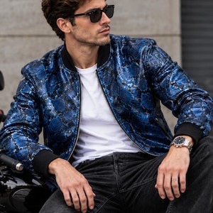 Könnte beinhalten: Eine blau-schwarze Bomberjacke mit Schlangenledermuster, Frontreißverschluss sowie schwarzem Rippstrickkragen und Rippstrickbündchen. Die Jacke wird über einem schlichten weißen T-Shirt getragen und mit einer dunkelgrauen Jeans sowie einer Sonnenbrille.