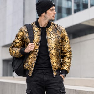 Peut inclure: Un blouson bomber à motif peau de serpent doré et noir avec une fermeture éclair frontale, un col, des poignets et un ourlet côtelés noirs. Le blouson est porté sur un t-shirt noir uni, avec un pantalon cargo noir, un bonnet et un sac à dos.