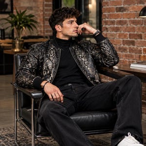 May include: A black and gold snakeskin print bomber jacket with a ribbed collar and cuffs, worn over a black turtleneck sweater, paired with black denim jeans and white sneakers. The jacket features a full-length front zipper and long sleeves.